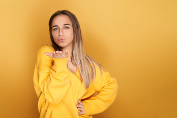 Fototapeta premium young girl in yellow sweater sends a kiss in the air, looking at camera. Isolated on yellow background.