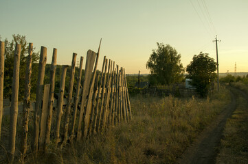 A dilapidated wooden fence along a rural road stretching into the distance, on a summer evening at sunset