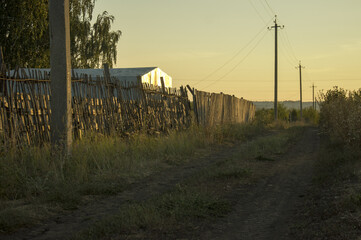 Rural summer evening landscape with a road, a dilapidated wooden fence and electric poles at sunset