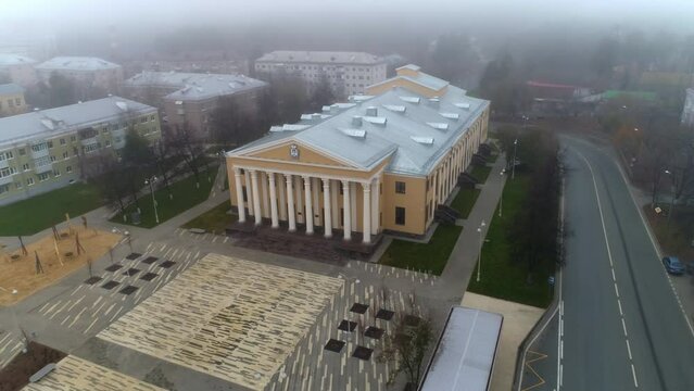 Large square with young trees near vintage palace of culture building next to road in autumn city on misty day aerial view
