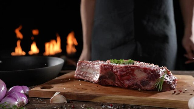 Raw seasoned rib eye steak meat beef dropped on wooden chopping board on a wooden table prepared for cooking with flames in the background. slow motion