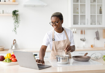 Fototapeta na wymiar Smiling millennial african american female in glasses, apron cooking, watching video lesson on laptop