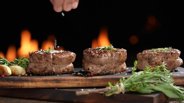 Cooked juicy steak meat beef with hand sprinking seasoning on top on wooden chopping board with flames in the background. slow motion