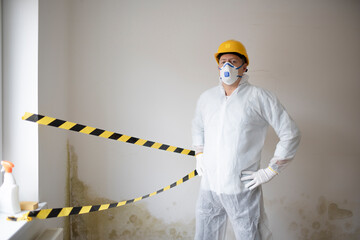 Man with protective mask, protective suit and yellow safety helmet tries to remove mold on wall...