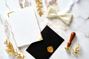 Wedding stationery set top view. Blank paper card mockup, black envelope, butterfly tie, wax stamp, golden branches on marble table.