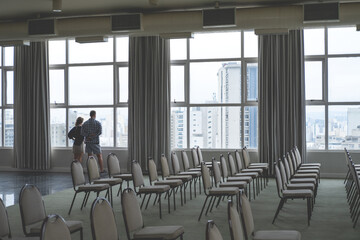 Two people a man and a woman look out a large window in an empty conference room with rows of empty chairs