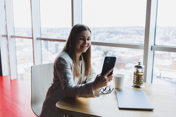 Obraz premium Cheerful young woman laughing while reading funny notification on smart phone sitting front laptop in cafe. Happy woman sending money through internet banking app on mobile phone