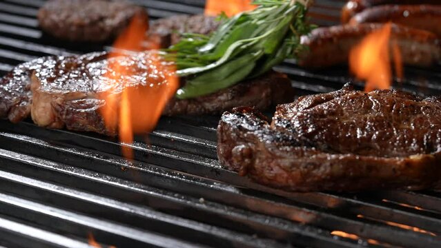 Cooked juicy steak meat beef brushed with rosemary on a flaming grill surrounded by other meat and sausages. slow motion