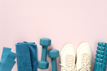 Feminine workout equipment top view. Dumbbells, fitness tape, towel, female sneakers, roller on pastel pink table.