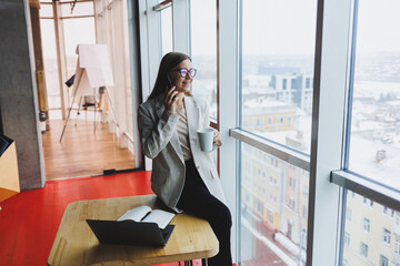 Fototapeta premium Young attractive female manager is talking on a mobile phone and smiling while sitting alone near a large window in a cafe in her free time and working on a computer. Happy woman is resting in a cafe.