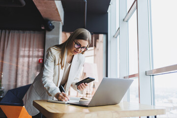 Young attractive female manager is talking on a mobile phone and smiling while sitting alone near a large window in a cafe in her free time and working on a computer. Happy woman is resting in a cafe.