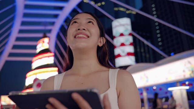 at night,young Asian woman happily plays with the tablet .She looks forward with hopes and dreams, a technology device of today in the middle of an urban society with buildings a in the backdrop.
