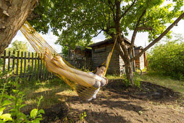 Village photo in the form of a girl in a hammock stretched between two apple trees and a bicycle near a wooden shed in the background. Rustic authentic portrait. Rural landscape.