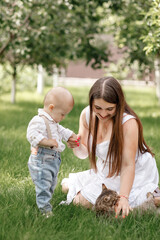 beautiful mother and her baby son playing with striped cat in the sunny summer park. mothers, baby's day. happy summer holiday