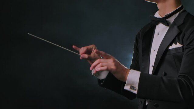 Concert of classical music under direction of an orchestra conductor. Man waves his baton to synchronize and conducting musicians in theater. Black background with smoke and backlight. Close up.