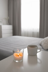 Burning candle and cup of tea or coffee stand on the bedside table. Bedroom in grey colors.