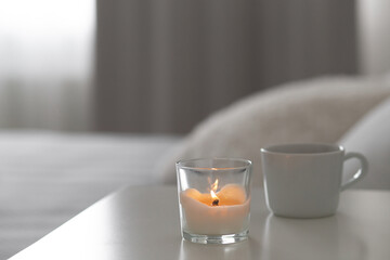 Burning candle and cup of tea or coffee stand on the bedside table.