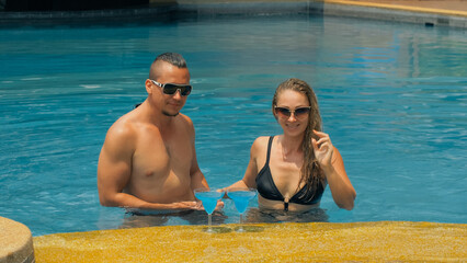 Fototapeta premium The loving couple hugs and kisses, drinking blue cocktail alcohol liquor in swimming pool at hotel outdoor. Portrait of caucasian man and woman. Creative hairstyles bodybuilder, swimsuit, sunglasses.
