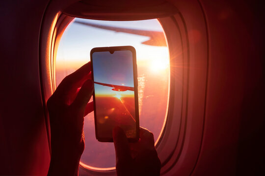 Photographing on a smartphone through the window of an airplane. The passenger's hands are holding the phone. View of the wing of the plane and the sunset. Selective focus. Close-up.