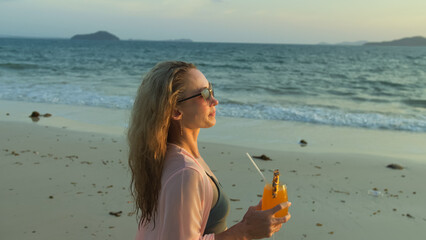 Fototapeta na wymiar Pretty woman walks on golden sunset on tropical beach against background of sea. Girl in green swimsuit and pink tunic silk shirt cape, drinks her orange cocktail Pina Colada with slice of pineapple