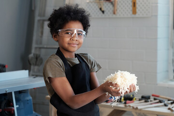 Waist up portrait of cute black boy holding wood shavings and looking at camera while enjoying work...