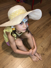 girl in a straw hat is sitting on the wooden floor hugging her knees and her eyes open and stare.