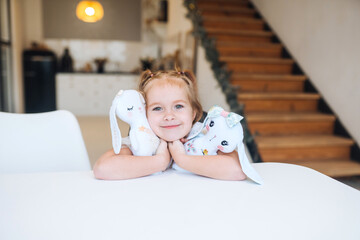 Little smiling girl hugging favorite toys while sitting at table