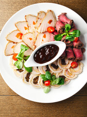 Antipasto catering platter on wooden background. Various types of meat, pork, beef, cherry, salami, rol with prunes, ham, basil, sauce, laid out on a wooden board close up. Space for text.
