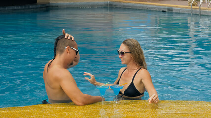 The loving couple hugs and kisses, drinking blue cocktail alcohol liquor in swimming pool at hotel outdoor. Portrait of caucasian man and woman. Creative hairstyles bodybuilder, swimsuit, sunglasses.