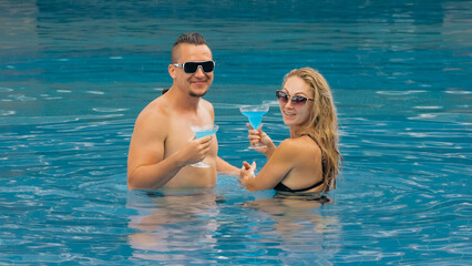 The loving couple hugs and kisses, drinking blue cocktail alcohol liquor in swimming pool at hotel outdoor. Portrait of caucasian man and woman. Creative hairstyles bodybuilder, swimsuit, sunglasses.