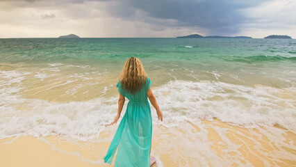 Beautiful long-haired woman walks to the stormy cloudy ocean on sand beach. Girl in blue swimsuit dress tunic. Concept rest in sea, tropical resort coastline traveling tourism summer beach holidays