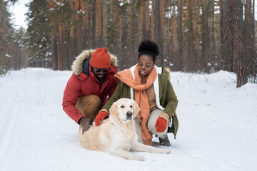 African young couple in warm clothing touching the dog lying on the snow during their walk in the...