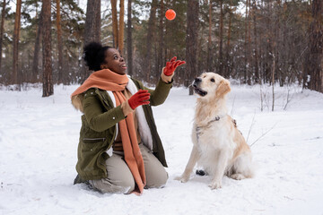 African woman in warm clothing playing ball with her dog during their walking in the forest in...