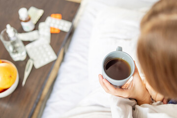 Sick woman covered with blanket holding cup of hot tea sitting on the bed. Young woman is treated at home, took sick leave