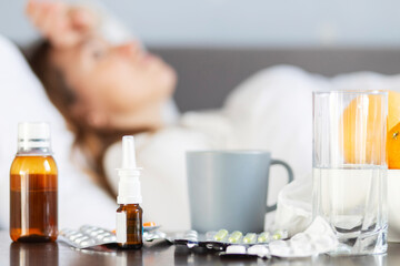 Sick young woman covered with a blanket lying in bed with high fever and a flu. A lot of medicines on table in bedroom