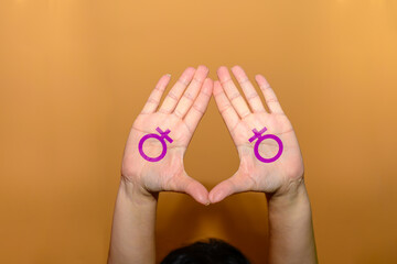 Two Caucasian woman's hands forming a triangle and painted with the female symbol in purple on a yellow background.