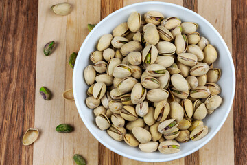 A white bowl is filled with pistachios.  Addional pistachios scrattered to left side.  Butcher block wood background.