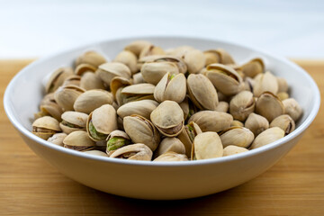 A white bowl of pistachios on a bamboo board with a white background.