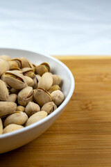 A white bowl of pistachios on a bamboo board with a white background.  Copy space.