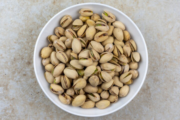 Top down view of a white bowl of pistachios on a tile surface.