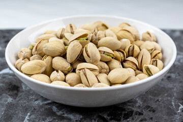 Side view of a white bowl of pistachios on a black marble surface.  White background.