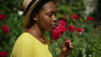 Side view young slim woman smelling fragrance of red rose in slow motion looking up at sunshine. Portrait of confident romantic African American gardener with flower in sunbeam outdoors in garden