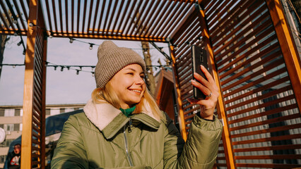 Young woman smiling doing selfie using smartphone at the city on a winter sunny day at a street food fair in an urban space.