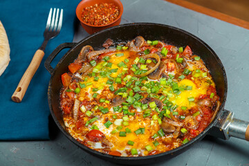 Shakshuka in a frying pan on a gray background next to vegetables and bread on a blue napkin.
