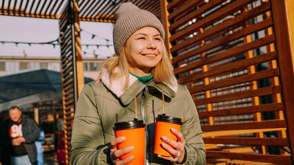 Woman carries two cups of mulled wine at the city outside food court. Street food festival, place to meet friends. She holds the cups forward.