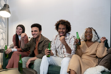 Excited young people sitting on sofa and watching sport match while drinking beer during home party