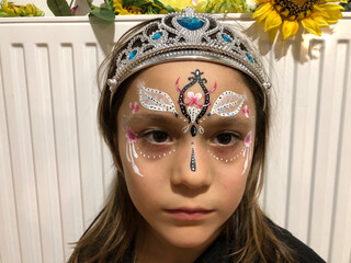 little sad girl with a tiara on her head on the white background and flowers. princess with a art face pattern  look away