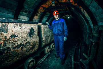 A tired miner in an old coal mine is standing near a broken trolley. A miner in a blue protective suit with an orange helmet on his head. Copy space.