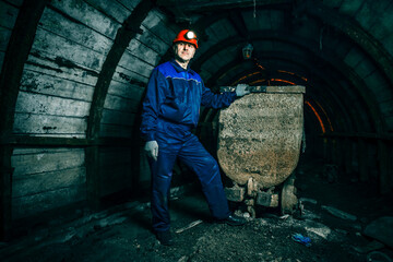 A tired miner in an old coal mine is standing near a broken trolley. A miner in a blue protective...