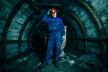 A tired miner in an old coal mine fixes a lantern. A miner in a blue protective suit with an orange...